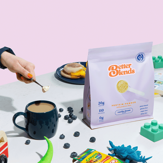 Better Blends Vanilla Dream protein powder bag on a table with a spoon of powder being added to coffee, surrounded by blueberries, toys, and breakfast items.