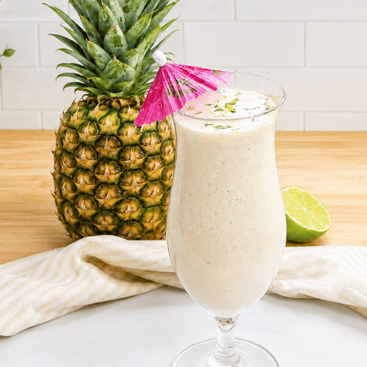 Creamy piña colada-inspired protein smoothie topped with fresh lime zest and a pink umbrella, served beside a whole pineapple and lime.