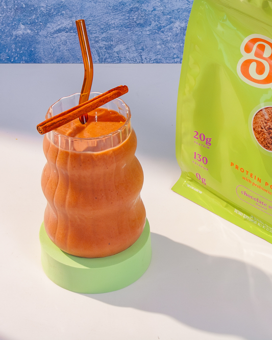 glass containing brown shake topped with cinnamon stick. A green bag sits to the right of the glass cup. 