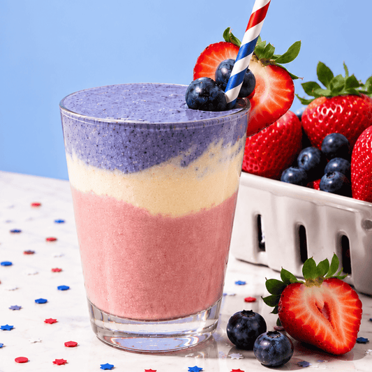 Red, white, and blue layered protein smoothie made with strawberries, vanilla yogurt, and blueberries, topped with fresh berries and a patriotic striped straw.