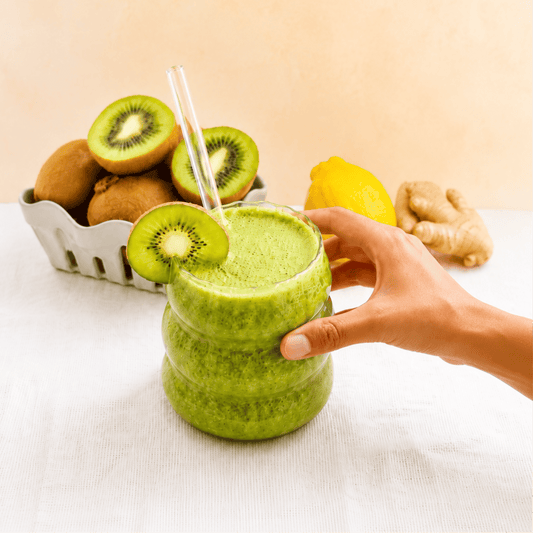 A refreshing green kiwi smoothie in a clear glass with a reusable glass straw, garnished with a fresh kiwi slice. A hand holds the glass while whole and halved kiwis, a lemon, and fresh ginger sit in the background on a light tabletop, creating a bright, 