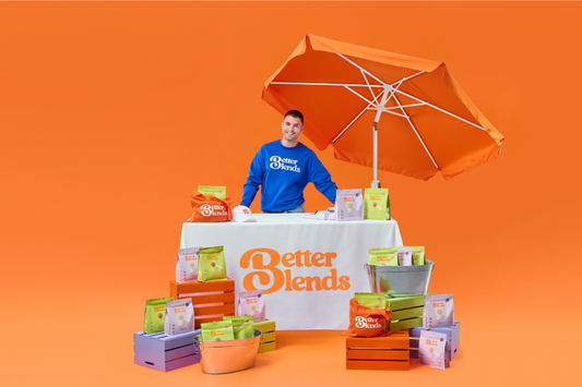 man in blue sweatshirt stands behind a table sourounded by bag of Better Blends protein powder. 