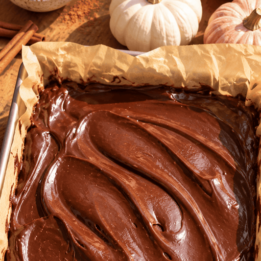 No-bake chocolate pumpkin protein brownies topped with rich chocolate ganache, served in a parchment-lined baking dish with pumpkins and cinnamon sticks in the background.